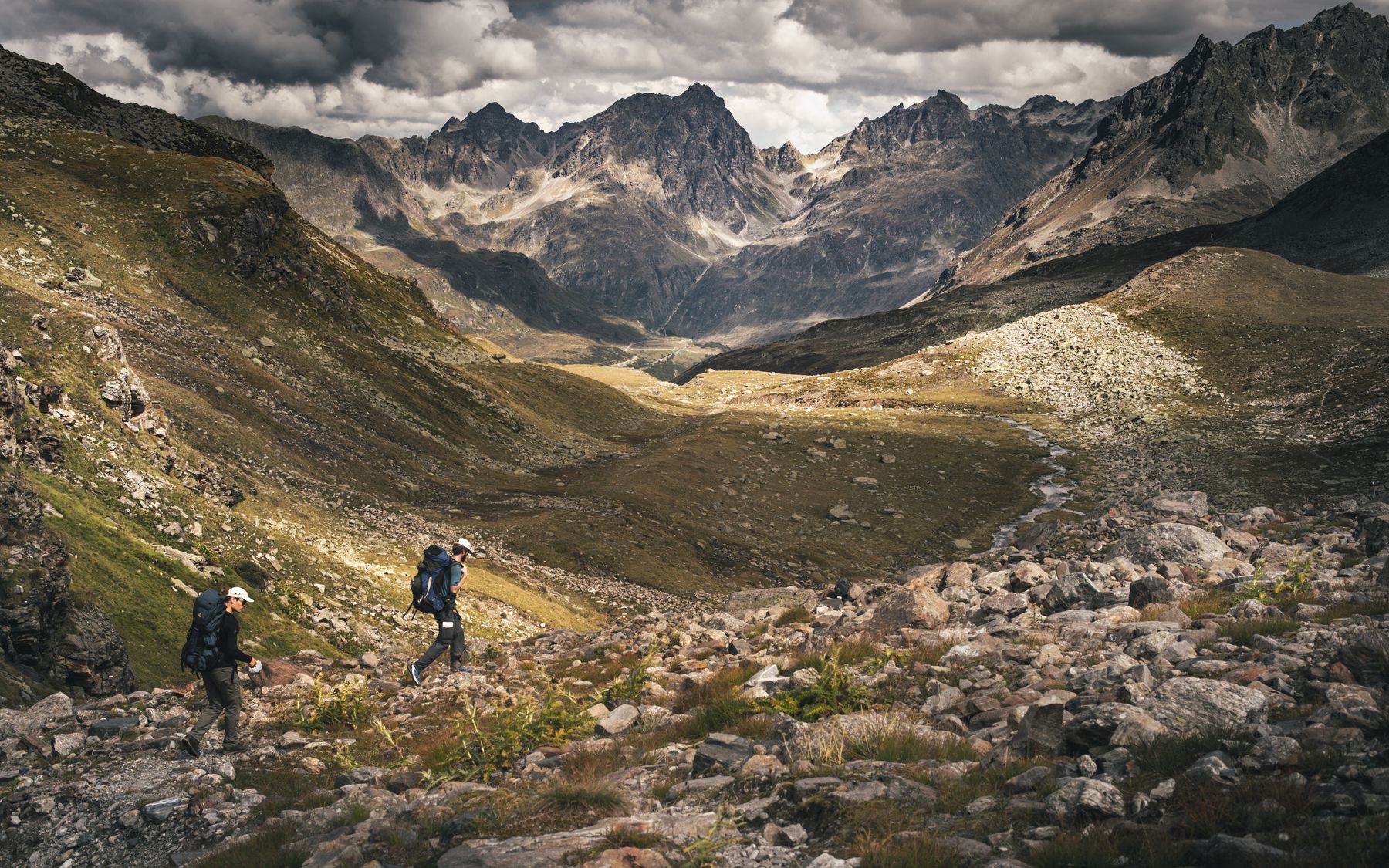 Two hikers explore rocky trails of the Austrian Alps under a dramatic cloudy sky